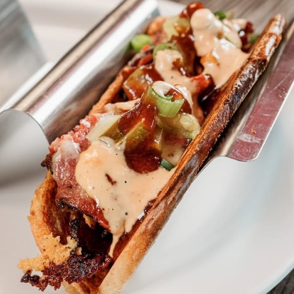 Bone Ends' BBQ hot dog with sauce, pickles, and green onions in a metal holder.